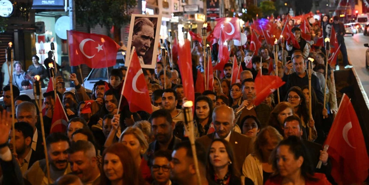 Akşam saatlerinde kalabalık bir grup insanın ellerinde Türk bayrakları ve meşalelerle gerçekleştirdiği 29 Ekim Cumhuriyet Bayramı yürüyüşü. Kalabalığın ön sıralarında, üzerinde Mustafa Kemal Atatürk'ün portresinin bulunduğu bir çerçeveyi taşıyan kişiler yer alıyor. İnsanlar neşeli ve coşkulu bir ifadeyle caddede ilerlerken, arka planda şehir ışıkları ve çevre binalar görülüyor.