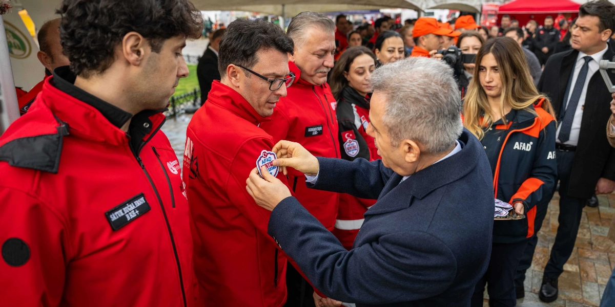 Koyu renk palto giymiş bir yetkili, kırmızı AFAD üniformalı bir görevlinin koluna AFAD arması takarken, çevresinde diğer AFAD personeli ve resmi yetkililer bulunuyor.
