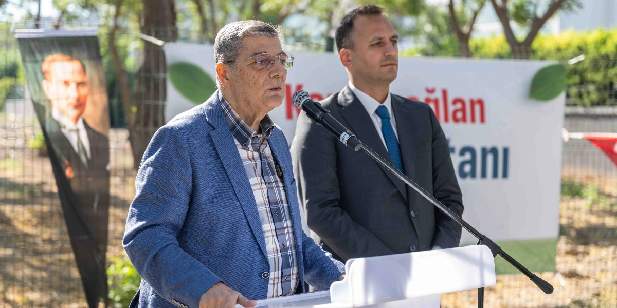 Bornova Belediye Başkanı Mustafa İduğ, kürsüde konuşma yaparken yanında bir yetkiliyle birlikte duruyor. Arka planda üzerinde 