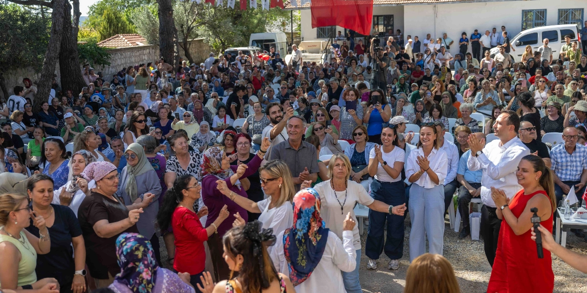 Açık havada düzenlenen Bornova Reçel Şenliği'nde, kalabalık bir katılımcı grubu müzik eşliğinde dans ediyor. Ön planda, ellerini havaya kaldırarak oyun oynayan kadınlar ve onları izleyip alkışlayan katılımcılar yer alıyor. Arka planda ise ağaçların altında oturarak şenliği takip eden çok sayıda kişi ve beyaz bir minibüs görülüyor. Ortamda şenlik havası hakim.