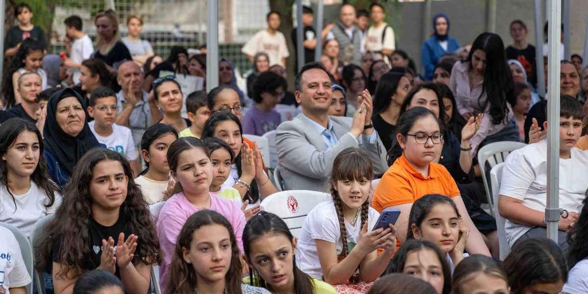 Açık havada düzenlenen bir etkinlikte, sandalyelerde oturan ve dikkatle sahneye bakan kalabalık bir öğrenci grubu ve yetişkinler. Ön planda, elleriyle alkışlayan veya dikkatle izleyen çeşitli yaşlarda öğrenciler yer alırken, orta kısımda açık renkli ceket giymiş bir erkek katılımcı alkışlıyor. Arka planda ise ayakta duran veya oturan çok sayıda izleyici bulunuyor.