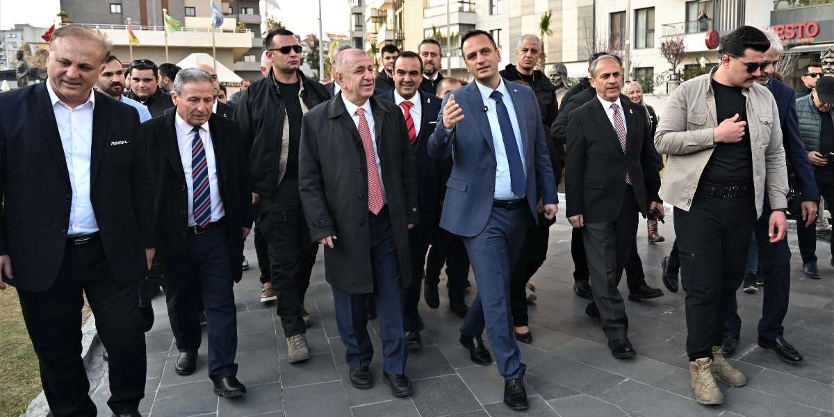 Ümit Özdağ ve beraberindeki bir grup erkek, Abide-i Hürriyet Meydanı'nda yürürken görülüyor. Özdağ, koyu renk bir palto ve kırmızı kravatla grubun ön sıralarında yer alırken, yanındaki mavi takım elbiseli bir kişi el hareketleriyle bir şeyler anlatıyor. Grubun geri kalanı takım elbiseli ve resmi kıyafetli kişilerden oluşuyor, arka planda ise modern binalar ve meydanın çevre düzenlemesi yer alıyor.