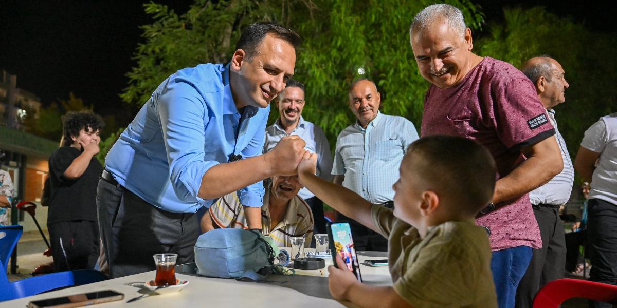 Açık mavi gömlekli bir erkek, gece vakti dışarıdaki bir masada oturan küçük bir çocukla yumruk tokuşturarak selamlaşıyor. Çevredeki diğer yetişkinler gülümseyerek bu anı izliyor, masada çay bardakları ve bir akıllı telefon bulunuyor.
