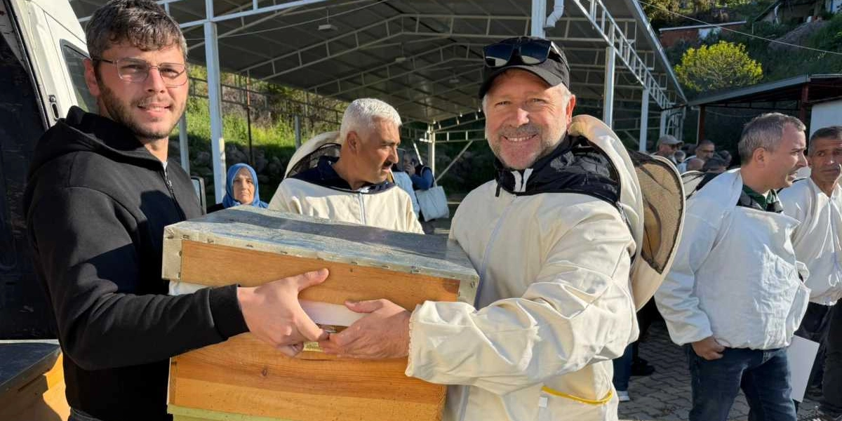 Açık havada, arıcılık kıyafeti giymiş bir adam ve yanında siyah sweatshirtlü bir genç, birlikte ahşap bir arı kovanını tutarak gülümsüyor. Arka planda arıcılık ekipmanları giymiş diğer insanlar ve metal çatılı bir yapı bulunuyor.