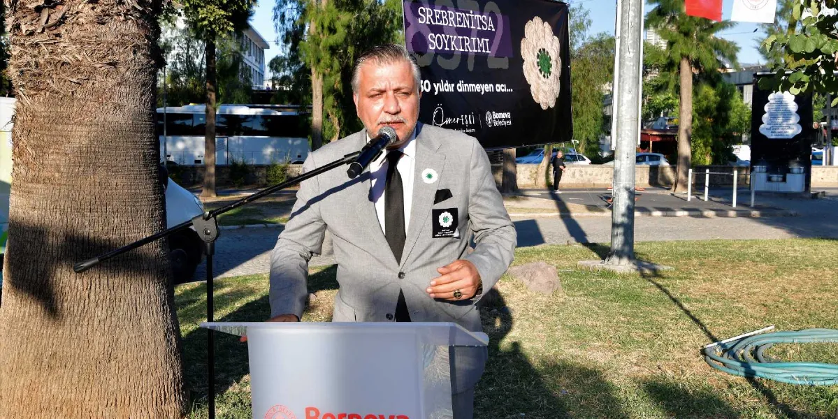 Gri takım elbiseli ve siyah kravatlı bir erkek, açık havada bir kürsüde konuşma yapıyor. Yaka kısmında Srebrenitsa'yı simgeleyen 