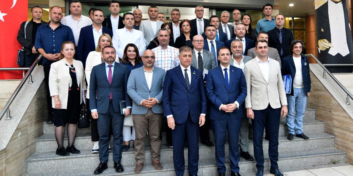 Bir binanın girişindeki merdivenlerde yan yana ve arka arkaya duran, çoğunluğu takım elbiseli erkeklerden ve birkaç kadından oluşan kalabalık bir grup. Ön sırada yer alan kişiler resmi kıyafetleriyle doğrudan kameraya bakarken, arka sıralardakiler daha rahat bir duruş sergiliyor. Sol tarafta Türk bayrağının bir kısmı, sağ tarafta ise üzerinde Atatürk silüeti bulunan bir afişin kenarı görülüyor.