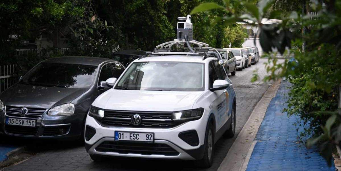 Tavanında özel bir haritalama ve görüntüleme cihazı bulunan beyaz bir Volkswagen marka araç, ağaçlıklı bir sokakta park halindeki diğer araçların yanında ilerliyor. Aracın yan kapısında kurumsal logolar ve yazılar yer alıyor.