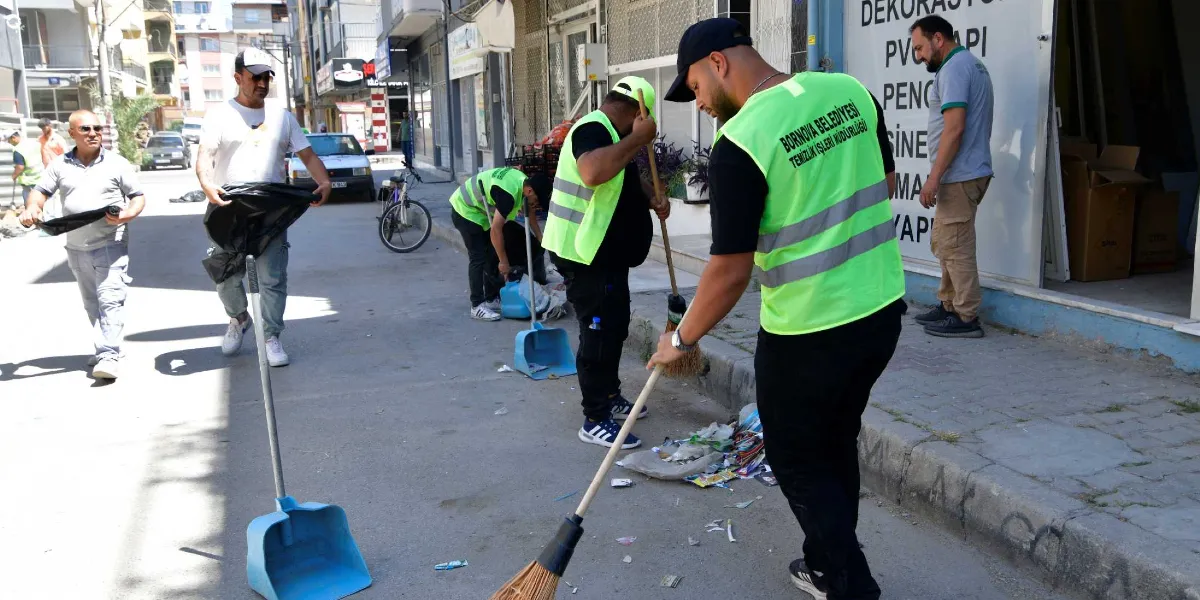 Bornova Belediyesi Temizlik İşleri Müdürlüğü yelekleri giymiş bir grup çalışan, bir sokakta süpürge ve faraşlarla temizlik yapıyor. Ön planda yer alan görevli sokağı süpürürken, diğer çalışanlar da çevredeki çöpleri topluyor. Arka planda dükkanların bulunduğu bir cadde ve park halindeki araçlar görülüyor.