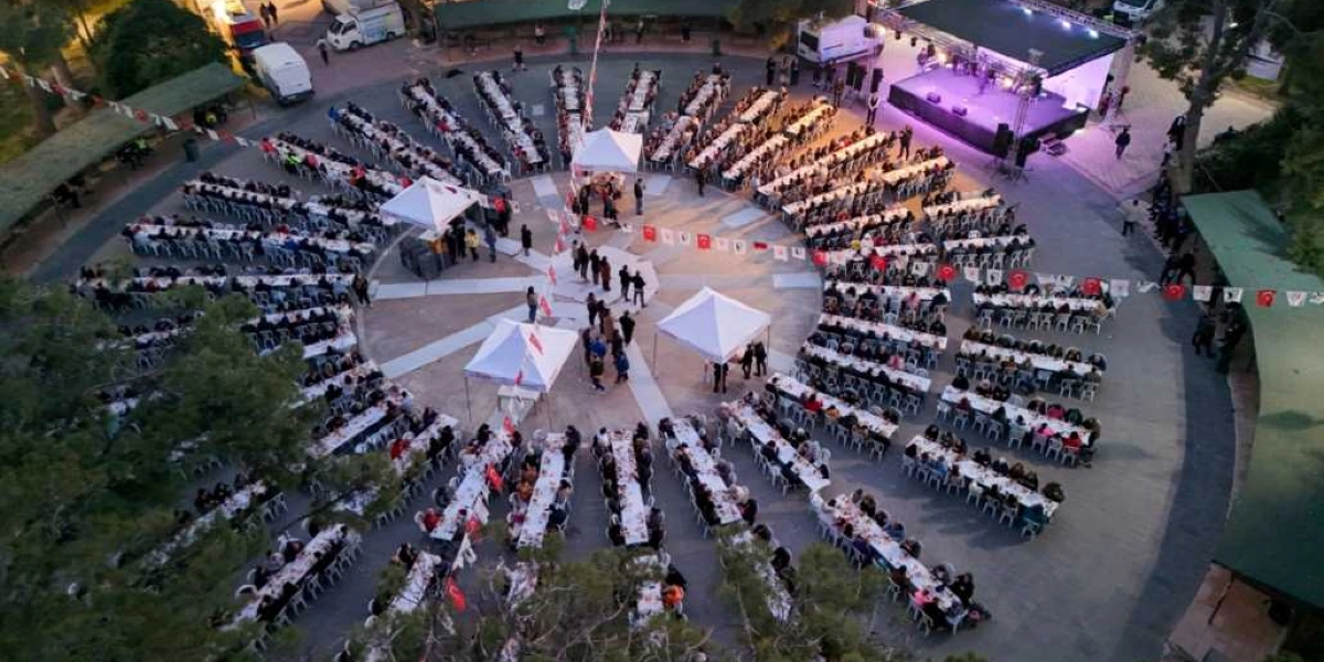 Açık havada, dairesel bir düzende yerleştirilmiş uzun masalarda oturan kalabalık bir grup insan, Kadir Gecesi iftar yemeği için bir araya gelmiş. Masaların merkezinde beyaz çadırlar bulunurken, alanın bir köşesinde ışıklandırılmış bir sahne yer alıyor. Etrafı ağaçlarla çevrili olan meydan, Türk bayraklarıyla süslenmiş.