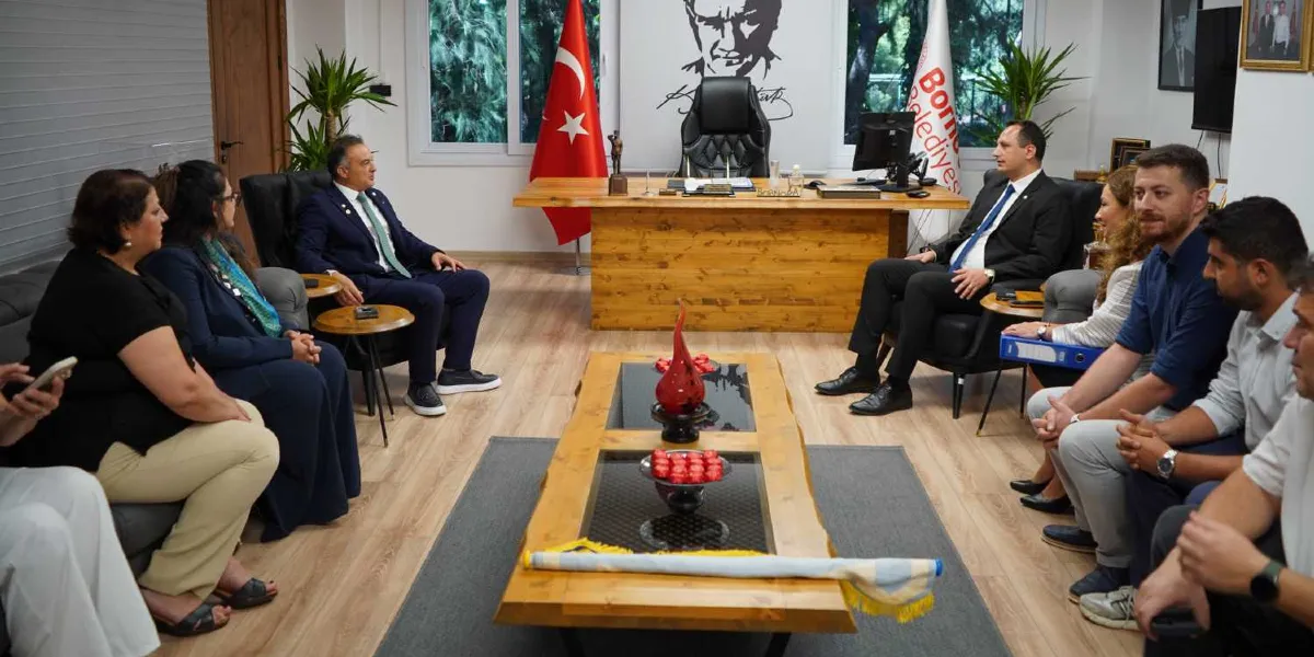 Modern bir ofis ortamında, karşılıklı koltuklarda oturan bir grup insan toplantı yapıyor. Odanın arka planında Türk bayrağı, Atatürk portresi ve üzerinde Bornova Belediyesi yazılı bir flama bulunuyor. Ahşap bir çalışma masasının arkasında oturan kişi ile karşısındaki katılımcılar, atık dönüşümü projesi üzerine görüşmelerini sürdürüyor. Ortadaki ahşap sehpanın üzerinde dekoratif kırmızı objeler ve bir flama yer alıyor.