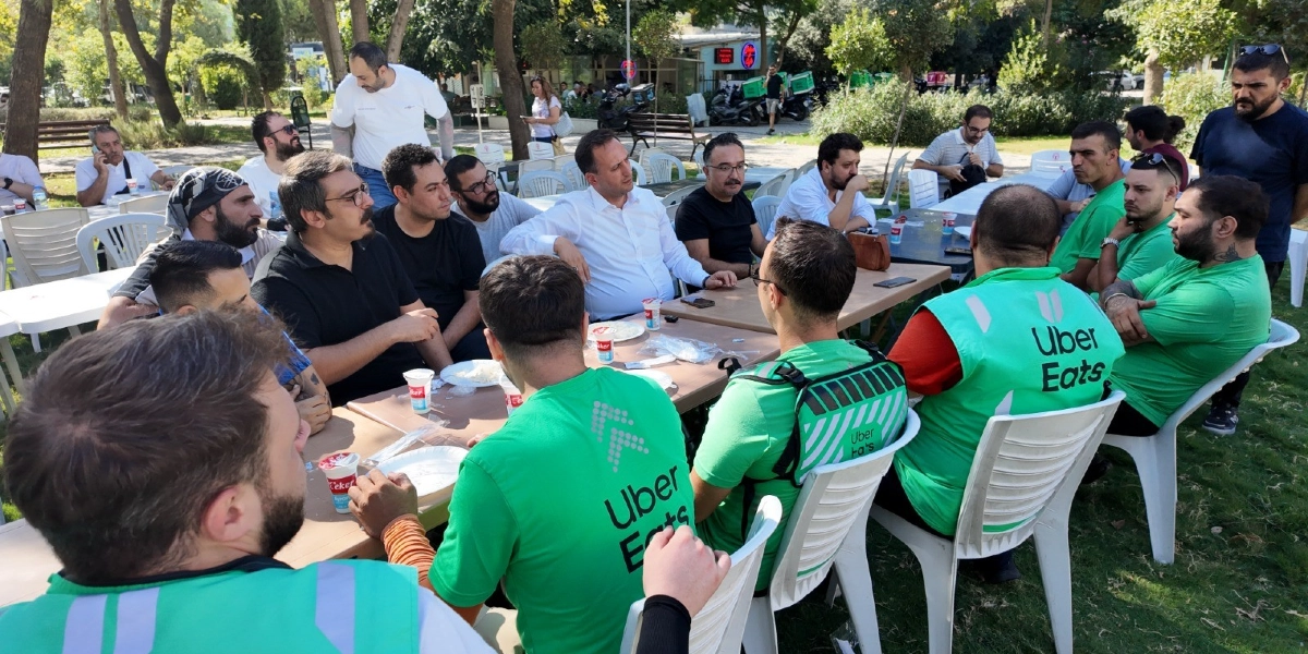 Açık havada, ağaçlık bir alanda plastik masalar etrafında toplanmış bir grup moto kurye ve katılımcı. Yeşil renkli, üzerinde beyaz Uber Eats logosu bulunan yelekler giymiş kuryeler masalarda oturuyor. Bazı katılımcılar beyaz gömlek ve tişört gibi günlük kıyafetlerle masada yer alırken, kuryeler yemek yiyor ve sohbet ediyor. Arka planda park ortamı, ağaçlar ve park halindeki motosikletler görülüyor.