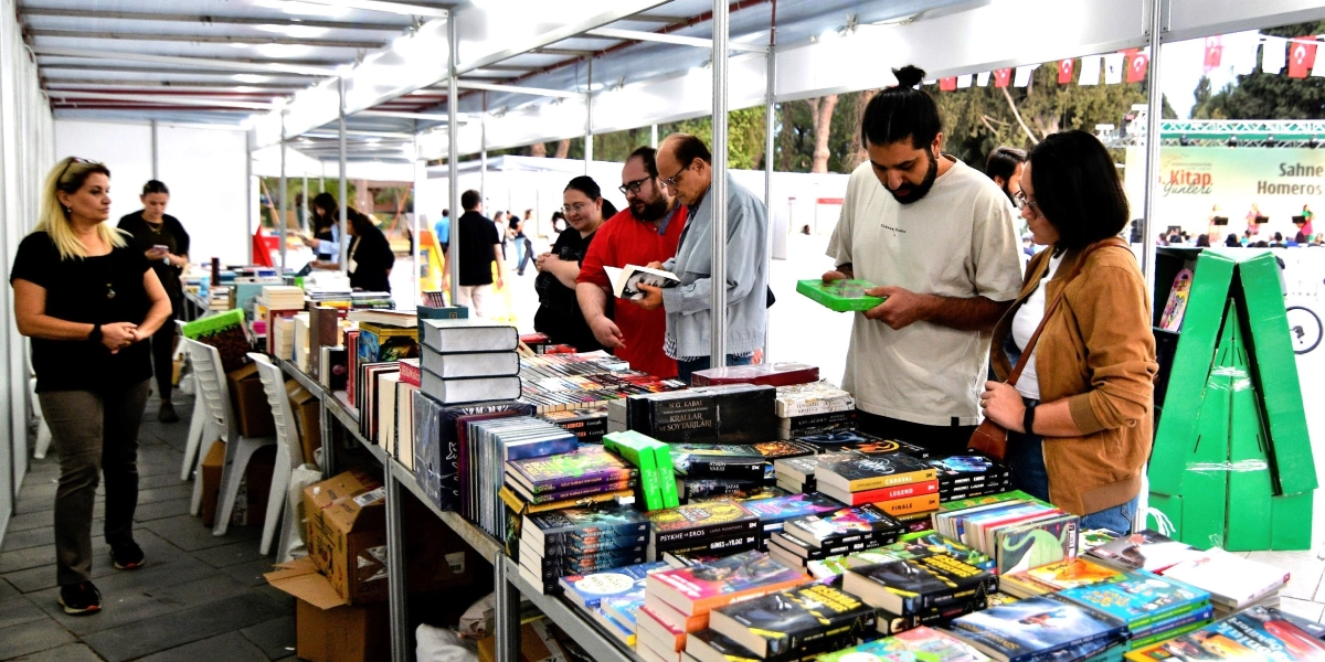 Bornova Kitap Günleri kapsamında kurulan bir stantta, uzun bir masanın üzerine dizilmiş kitapları inceleyen ziyaretçiler görülüyor. Ön planda, elinde yeşil kapaklı bir kitap tutan sakallı bir genç adam ve yanında duran gözlüklü bir kadın kitaplara bakıyor. Arka planda ise farklı kitapları inceleyen diğer ziyaretçiler ve stant görevlileri yer alıyor. Stant alanının arka tarafında, üzerinde 