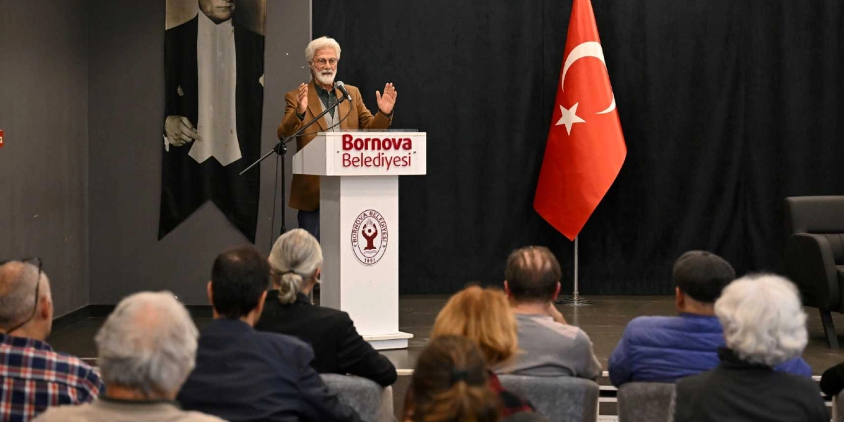 Beyaz saçlı ve sakallı bir konuşmacı, üzerinde Bornova Belediyesi logosu bulunan bir kürsüde, ellerini kullanarak dinleyicilere hitap ediyor. Konuşmacının arkasında sol tarafta Mustafa Kemal Atatürk'ün bir portresi, sağ tarafta ise Türk bayrağı yer alıyor. Salonun ön kısmında, konuşmacıyı dinleyen bir grup katılımcı arkaları dönük şekilde oturuyor.