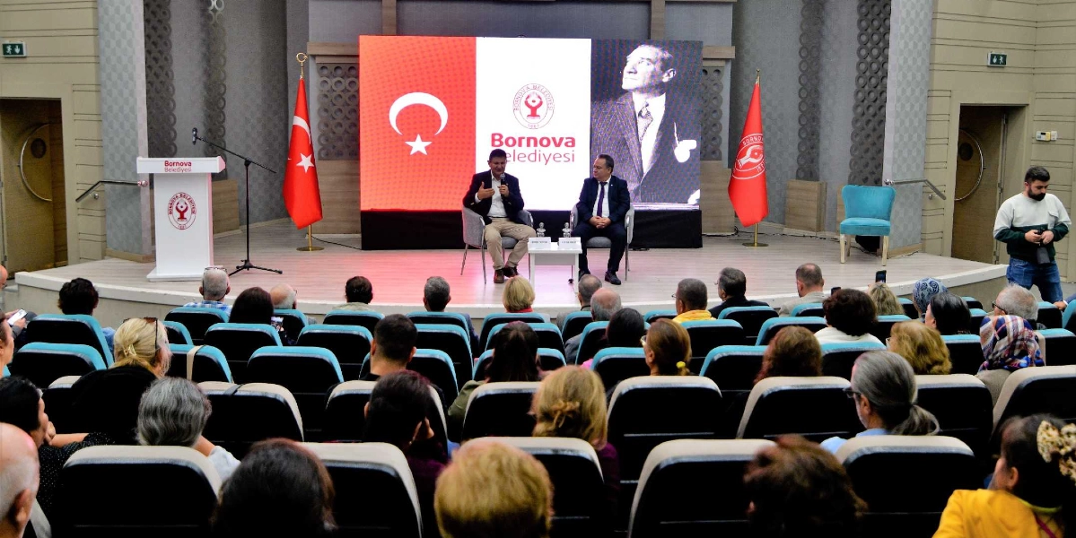 Bornova Belediyesi tarafından düzenlenen bir etkinlikte, sahnedeki iki konuşmacı izleyicilere hitap ediyor. Sahnenin arka planındaki dev ekranda Türk bayrağı, Bornova Belediyesi logosu ve Mustafa Kemal Atatürk’ün bir portresi yer alıyor. Sahnenin iki yanında da Türk bayrağı ve Bornova Belediyesi flaması bulunuyor. Konferans salonunda oturan izleyiciler, sahnedeki konuşmacıları dinliyor.