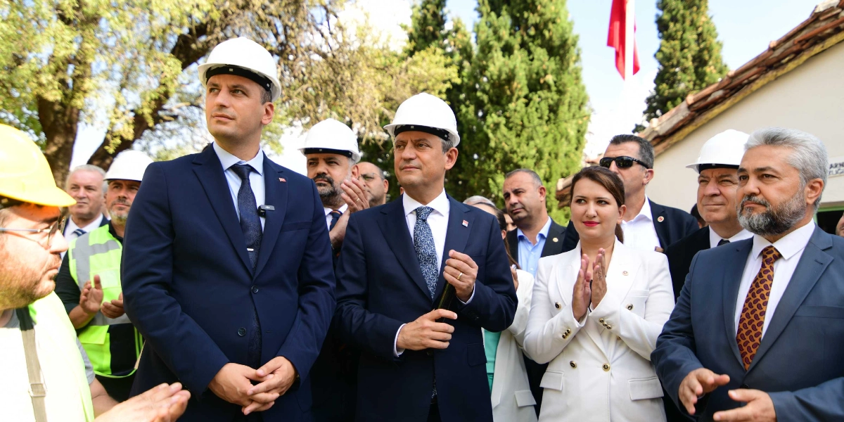 Özgür Özel ve beraberindeki heyet, başlarında beyaz inşaat baretleriyle bir açılış töreninde yer alıyor. Özel, lacivert takım elbisesi ve desenli kravatıyla ön planda dururken, yanında beyaz ceketli bir kadın ve diğer yetkililer bulunuyor. Arka planda ağaçlar ve Türk bayrağı görülürken, bazı katılımcılar alkışlıyor.