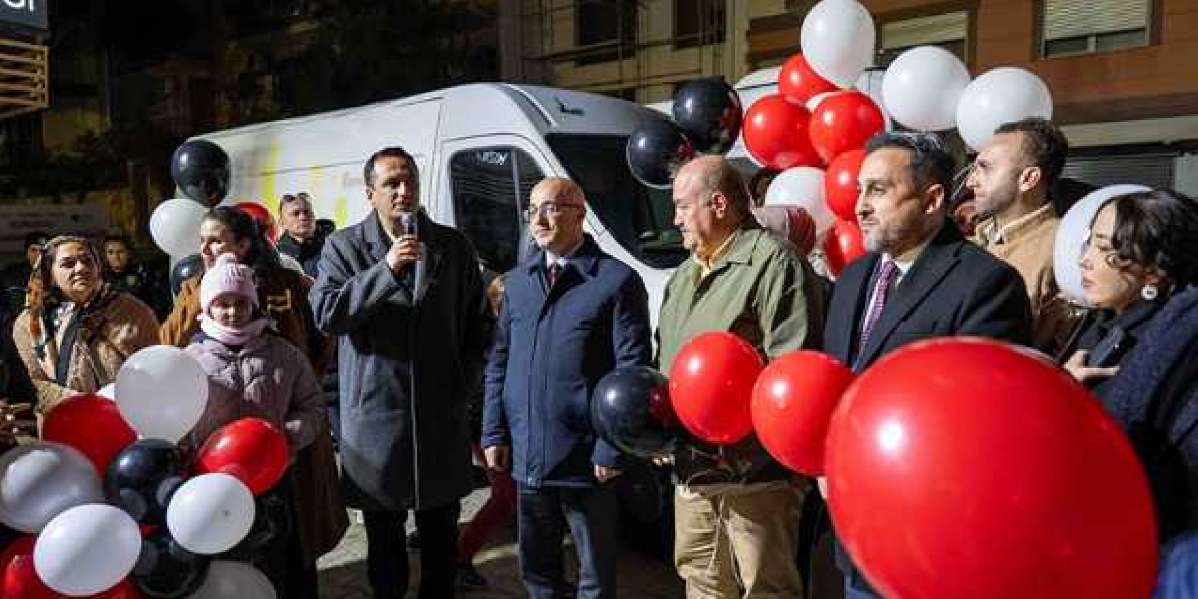 Gece vakti dış mekanda düzenlenen anma etkinliğinde, ellerinde kırmızı, beyaz ve siyah balonlar tutan bir grup insan yer alıyor. Gruptan bir kişi mikrofonla konuşma yaparken, yanındaki diğer katılımcılar onu dinliyor. Arka planda beyaz bir minibüs ve binalar görülüyor.