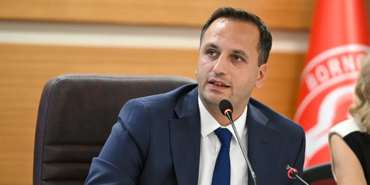 Lacivert takım elbiseli, beyaz gömlekli ve mavi kravatlı bir erkek, toplantı masasında önündeki mikrofona konuşuyor. Arka planda Bornova Belediyesi'ne ait olduğu anlaşılan kırmızı bir bayrak ve ahşap panelli bir duvar bulunuyor.