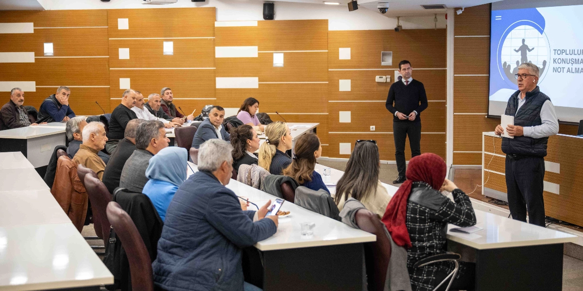 Modern bir konferans salonunda, sıralarda oturan bir grup muhtar ve katılımcı, ayakta sunum yapan bir konuşmacıyı dinliyor. Konuşmacı, elinde not defteriyle katılımcılara hitap ederken, yanında lacivert kazaklı başka bir yetkili ayakta duruyor. Salonun önündeki projeksiyon ekranında 