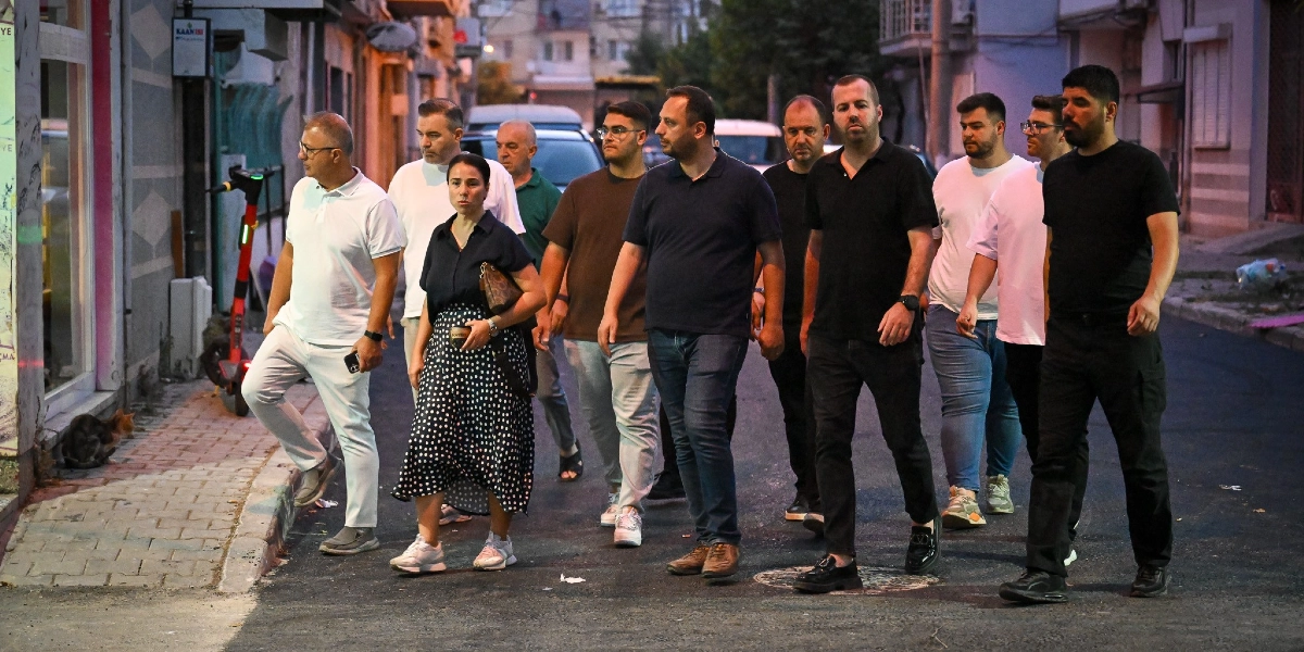 Çamdibi'nde akşam saatlerinde bir grup kadın ve erkek, asfaltlanmış bir sokakta yan yana yürüyerek incelemelerde bulunuyor. Grubun önünde beyaz tişörtlü ve beyaz pantolonlu bir erkek ile lacivert gömlekli ve puantiyeli etekli bir kadın yer alıyor; arkalarında ise çoğunluğu koyu renkli kıyafetler giymiş diğer grup üyeleri bulunuyor.