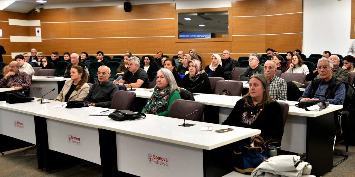 Bornova Belediyesi'ne ait bir konferans salonunda, sıralarda oturmuş dikkatle dinleyen kalabalık bir izleyici grubu. Beyaz masaların üzerinde Bornova Belediyesi logoları yer alıyor ve katılımcılar sahneye doğru dönerek toplantıyı takip ediyor.
