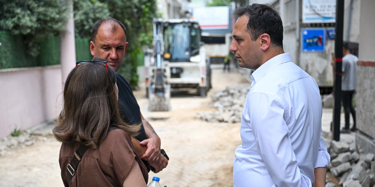 Bir sokak çalışması alanında, beyaz gömlekli bir erkek ile yanındaki kadın ve siyah tişörtlü bir erkek bir arada durarak konuşuyor. Arka planda ise inşaat çalışmaları için kullanılan bir iş makinesi ve kazılmış bir yol görülüyor.