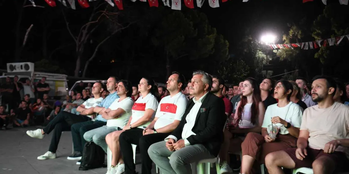 Gece vakti açık havada kurulan dev ekranda basketbol maçı izleyen kalabalık bir grup insan, sandalyelerde oturmuş dikkatle karşıya bakıyor. İzleyicilerin arasında Türk bayraklı tişörtler giymiş kişiler bulunuyor ve ortamda asılı Türk bayrakları yer alıyor.