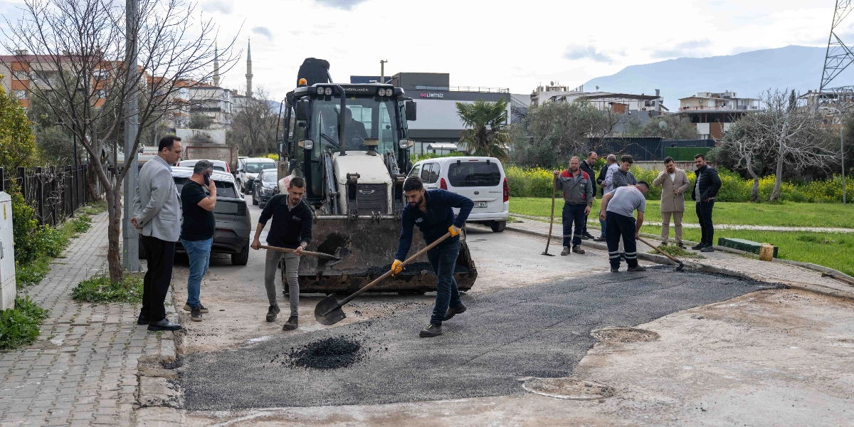 Bornova'da bir sokakta belediye ekipleri tarafından gerçekleştirilen yol yenileme çalışması. Bir iş makinesi asfalt dökümüne destek verirken, işçiler ellerindeki küreklerle asfaltı düzeltiyor. Çalışmaları izleyen yetkililer ve çevredeki araçlar görülüyor.