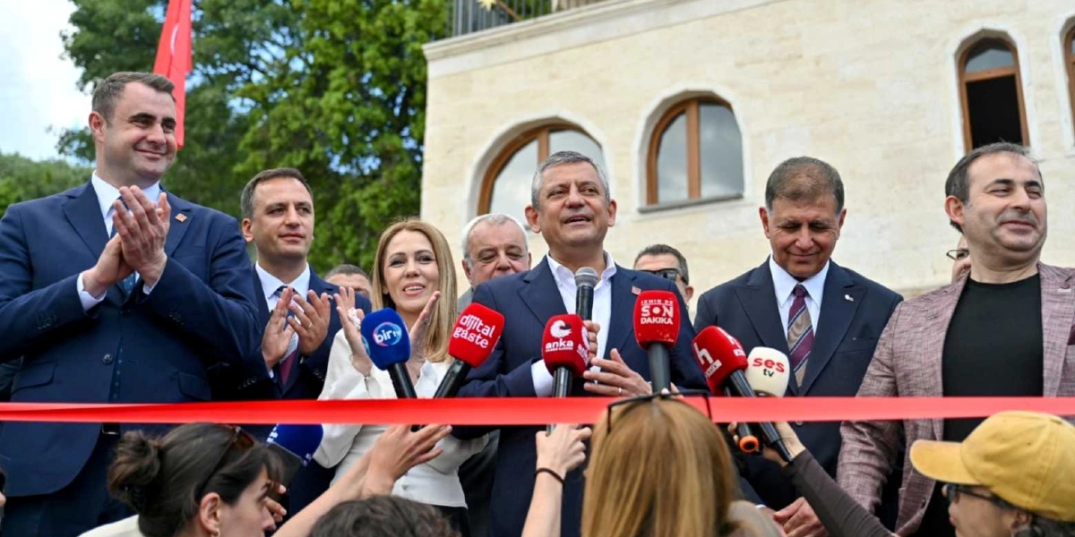CHP Genel Başkanı Özgür Özel, Bornova'daki bir açılış töreninde mikrofon başında konuşma yaparken, yanında yer alan diğer yetkililerle birlikte kırmızı kurdeleyi kesmeye hazırlanıyor. Özel'in önünde çeşitli haber kuruluşlarına ait mikrofonlar yer alıyor, çevresindeki katılımcılar ise alkışlarla kendisine eşlik ediyor. Arka planda ise tarihi dokuya sahip taş bir bina ve ağaçlar görülüyor.