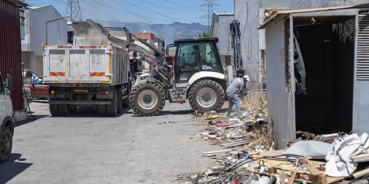 Bornova Belediyesi'ne ait bir iş makinesi, sokak üzerindeki ayakkabı ve diğer atıkları kepçesiyle toplayarak kamyonun kasasına yüklüyor. İş makinesinin yanında duran bir görevli, elindeki süpürgeyle atıkları temizlemeye yardımcı oluyor.
