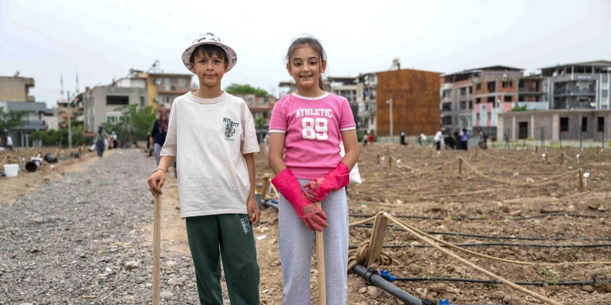 Kent bostanında ellerinde bahçe çapalarıyla duran, biri beyaz şapkalı ve beyaz tişörtlü, diğeri pembe tişörtlü ve pembe bahçe eldivenli iki çocuk, arka planda şehir yerleşimi ve ekim alanları ile birlikte kameraya gülümseyerek bakıyor.