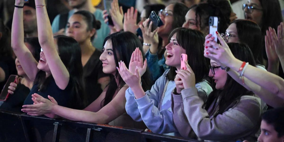 Bir konser alanında sahneye doğru dönmüş, heyecanla alkışlayan ve telefonlarıyla kayıt yapan gençlerden oluşan kalabalık bir grup. Ön planda yer alan genç kadınlar gülümseyerek sahnedeki performansı izliyor, bazıları ellerini havaya kaldırmış coşkuyla eşlik ediyor.