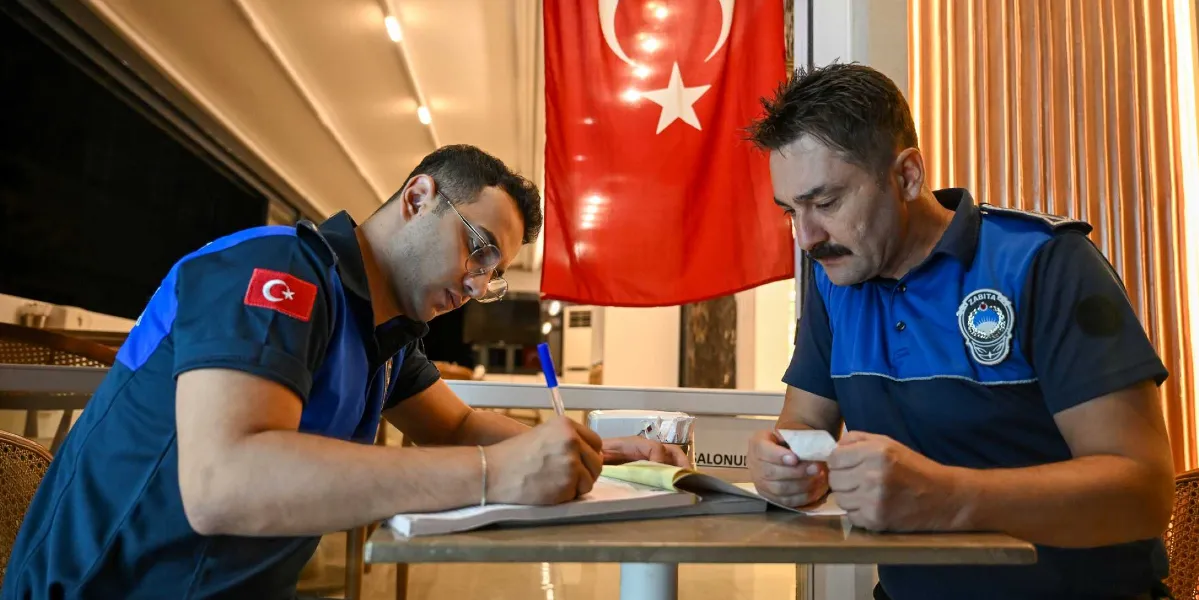 Mavi zabıta üniformalı iki görevli, bir masada oturarak denetim tutanağı tutuyor. Soldaki görevli elindeki kalemle evraka yazı yazarken, sağdaki görevli elindeki küçük bir kağıdı inceliyor. Arka planda duvarda asılı bir Türk bayrağı bulunuyor.