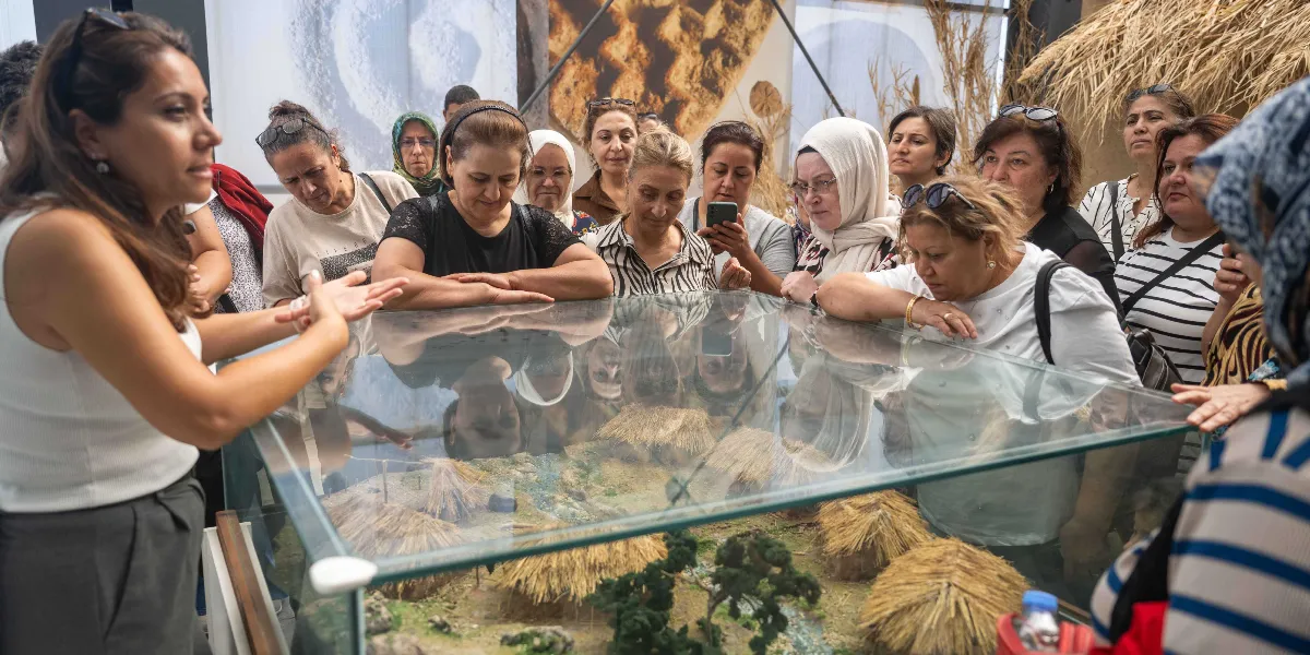 Bir rehber eşliğinde müze veya sergi alanında bulunan bir grup kadın, cam bir vitrin içerisinde sergilenen tarih öncesi bir yerleşim yerini temsil eden maketi dikkatle inceliyor. Rehber, ellerini kullanarak maket hakkında açıklamalarda bulunurken, ziyaretçiler camın etrafına toplanmış durumda. Makette sazdan yapılmış küçük kulübeler, ağaçlar ve bir dere yatağı detaylı bir şekilde yer alıyor.