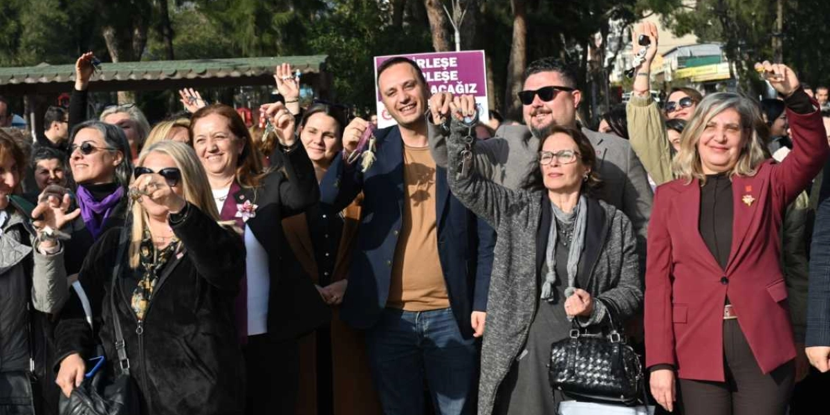 Açık havada, güneşli bir günde bir araya gelmiş kadın ve erkeklerden oluşan bir grup, ellerinde havaya kaldırdıkları anahtarlarla poz veriyor. Grubun arkasında üzerinde 