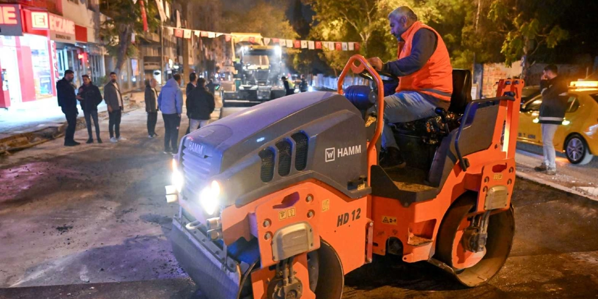 Gece vakti Bornova'da bir cadde üzerinde turuncu yelekli bir operatörün kullandığı Hamm marka silindir, asfalt serim çalışması yapıyor. Arka planda bir grup işçi ve görevli çalışmaları izlerken, yol kenarında park halindeki sarı bir taksi ve sol tarafta ışıklı bir eczane tabelası görülüyor.