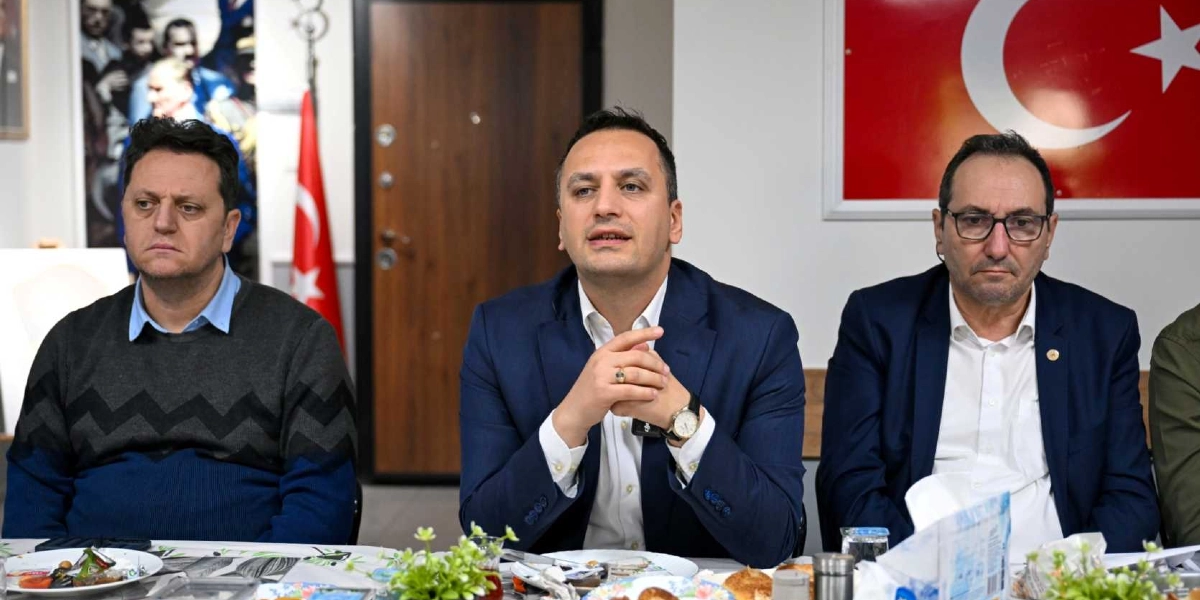 Bir masa etrafında yan yana oturan üç erkek, bir toplantı sırasında konuşma yapıyor. Ortada lacivert ceketli bir kişi ellerini birleştirmiş konuşurken, yanlarında gri kazaklı ve gözlüklü, lacivert ceketli diğer iki kişi onu dinliyor. Arka planda duvarda asılı bir Türk bayrağı ve Atatürk görseli bulunuyor.
