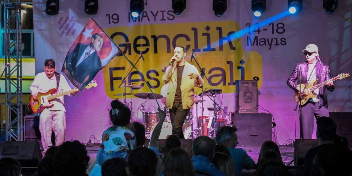 19 Mayıs Gençlik Festivali sahnesinde performans sergileyen üç kişilik bir müzik grubu. Ortada mikrofonla şarkı söyleyen solist, solda bas gitar çalan bir müzisyen ve sağda elektro gitar çalan güneş gözlüklü bir gitarist yer alıyor. Solist, üzerinde Atatürk portresi ve Türk bayrağı bulunan bir bayrağı havada tutuyor. Arka planda 14-18 Mayıs tarihli Gençlik Festivali yazılı bir afiş ve sahne ışıkları görülüyor. Ön planda ise konseri izleyen kalabalık bir izleyici kitlesi bulunuyor.