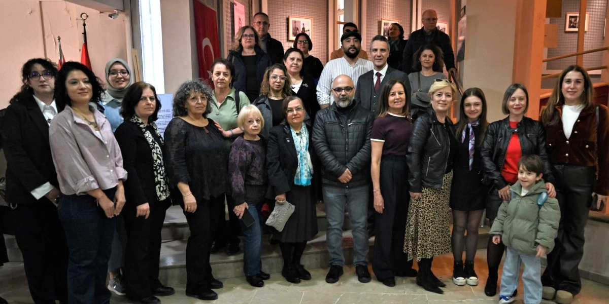 Kapalı bir mekanda, bir merdiven önünde yan yana duran, çoğunluğu kadınlardan oluşan kalabalık bir grup. Katılımcılar günlük ve şık kıyafetler içinde, gülümseyerek kameraya bakıyor. Grubun ön sırasında, sağ tarafta küçük bir erkek çocuğu da yer alıyor. Arka planda duvarda asılı Türk bayrağı ve çerçeveli fotoğraflar bulunuyor.