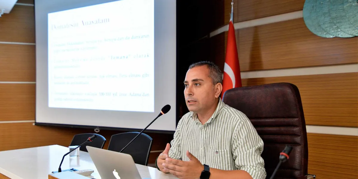 Toplantı salonunda, arkasında projeksiyon perdesi ve Türk bayrağı bulunan bir konuşmacı, önündeki mikrofon ve dizüstü bilgisayar eşliğinde sunum yapıyor. Konuşmacı, çizgili gömlek giymiş ve ellerini kullanarak bir şeyler anlatıyor; projeksiyon perdesinde ise 