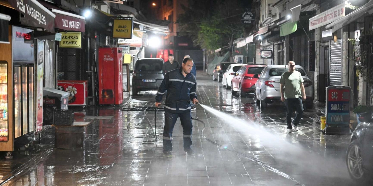 Gece vakti Bornova'da bir sokakta, üzerinde reflektörlü iş kıyafeti bulunan bir belediye temizlik görevlisi, elindeki basınçlı su hortumuyla ıslak zemini yıkıyor. Sokak boyunca dükkanlar, park halindeki araçlar ve yürüyen bir vatandaş görülüyor.