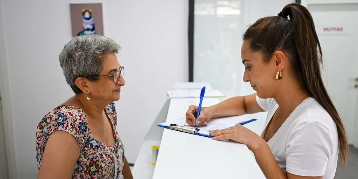 Eros Kedi Kliniği'nin resepsiyonunda, beyaz tişörtlü genç bir kadın görevli, elindeki dosyaya notlar alırken karşısında duran gözlüklü ve çiçek desenli bluz giyen orta yaşlı bir kadınla ilgileniyor. Arka plandaki duvarda kedi figürlü bir tablo asılı duruyor.