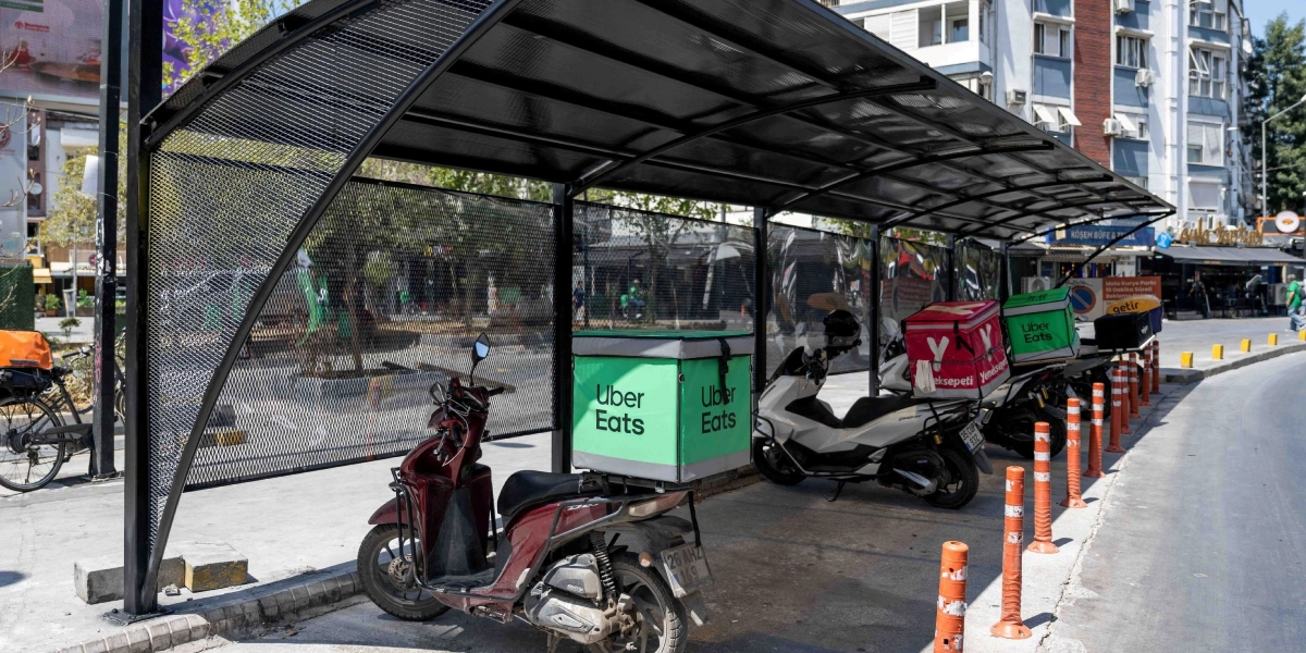 Bornova'da kuryeler için özel olarak tasarlanmış, güneş ve yağmurdan koruyan metal çatılı ve yanları ızgaralı bekleme durağı. Durağın altında park edilmiş, arkalarında Yemeksepeti, Uber Eats ve Getir gibi farklı yemek dağıtım firmalarının logolarını taşıyan termal çantaların bulunduğu motosikletler yer alıyor.