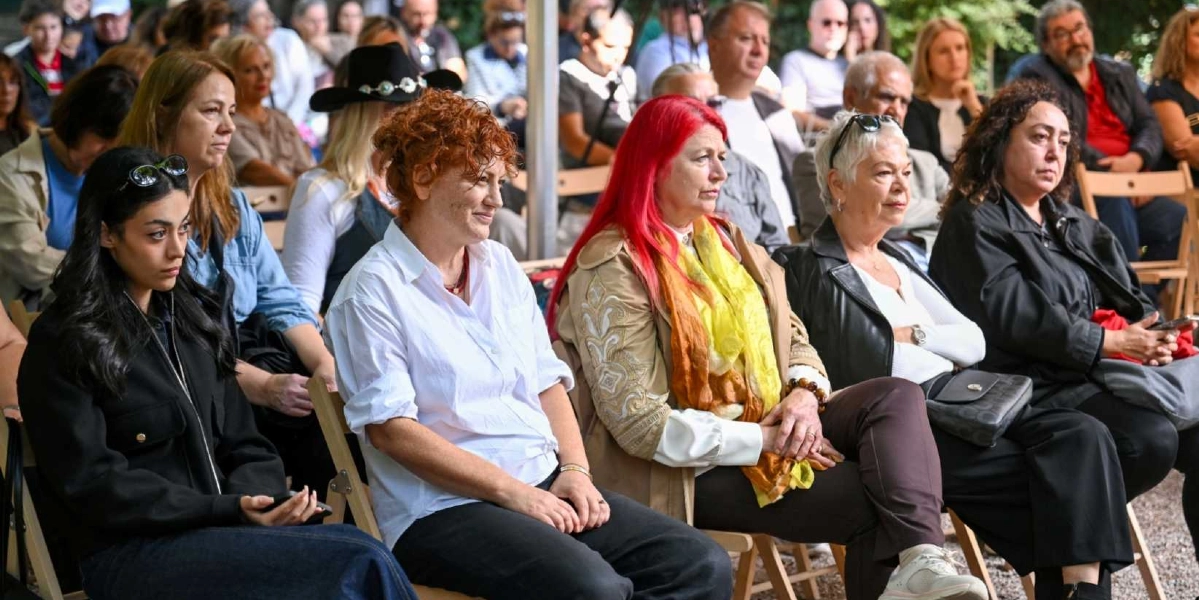 Açık havada düzenlenen bir etkinlikte yan yana oturmuş, dikkatle dinleyen bir grup kadın izleyici. Ön planda, siyah ceketli genç bir kadın, yanında beyaz gömlekli kızıl saçlı bir kadın, onun yanında parlak kırmızı saçlı ve sarı-turuncu şallı bir kadın, yanında kısa gri saçlı bir kadın ve en sağda siyah ceketli bir kadın yer alıyor. Arka planda ise etkinliği takip eden diğer katılımcılar görülüyor.