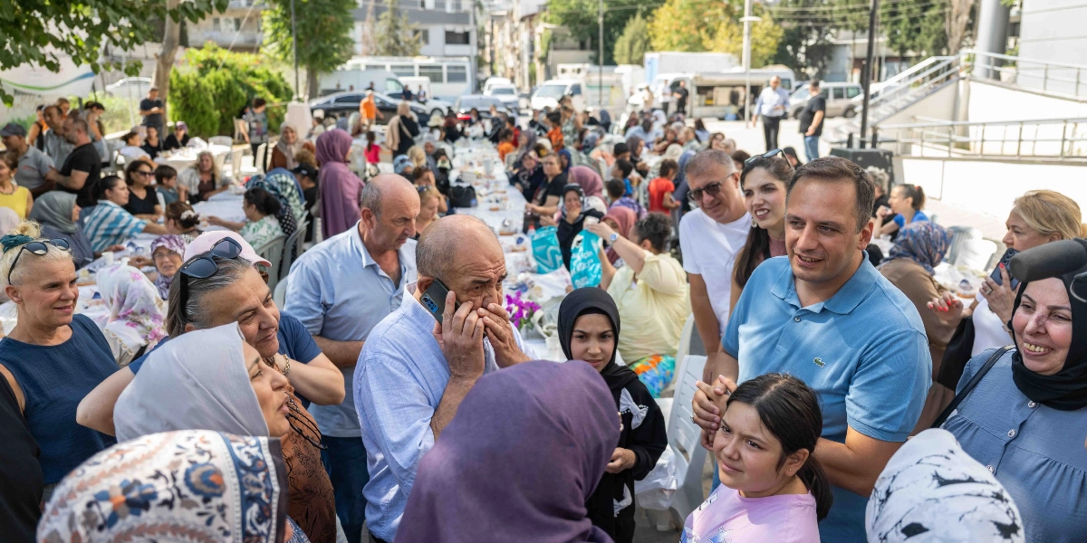 Altındağ Kent Bostanı'nda düzenlenen açık hava etkinliğinde, uzun masaların etrafında toplanmış kalabalık bir grup insan bir arada oturuyor. Ön planda, mavi polo yaka tişört giyen bir erkek, yanındaki genç kızla ve çevresindeki insanlarla sohbet ederken, hemen yanında telefonla konuşan yaşlı bir adam yer alıyor. Etrafı çeşitli yaş gruplarından kadınlar ve çocuklarla çevrili olan bu sosyal buluşmada, katılımcılar masalarda yemek yiyip keyifli vakit geçiriyor.