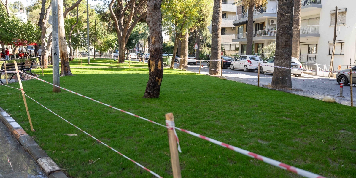 Büyükpark içerisinde, çevresi kırmızı beyaz güvenlik şeritleriyle çevrilmiş, yeni düzenlenmiş yeşil çim alan ve parkın hemen yanındaki yol kenarında park etmiş araçlar ile apartmanlar görülüyor.