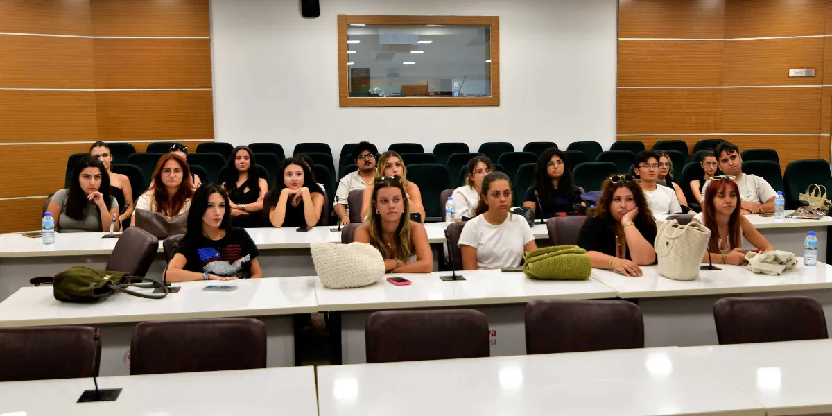 Modern bir konferans salonunda sıralarda oturan ve dikkatle karşıya bakan Peyzaj Mimarlığı öğrencileri. Öğrenciler beyaz masaların arkasında, koyu renkli sandalyelerde oturuyor; masaların üzerinde çantalar, su şişeleri ve telefonlar bulunuyor. Arka planda ahşap panellerle kaplı duvarlar ve ortada dikdörtgen bir pencere yer alıyor.
