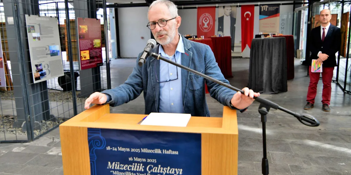 Gözlüklü, gri saçlı ve sakallı bir konuşmacı, üzerinde 