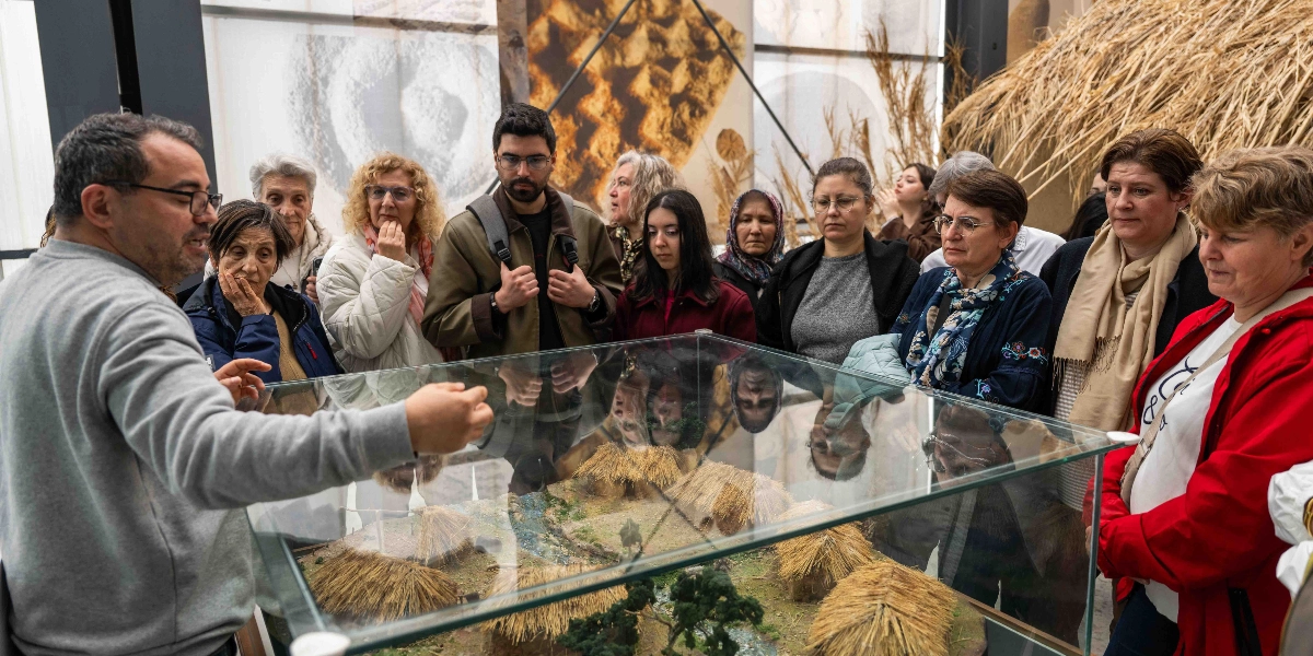Müze ortamında, gözlüklü bir rehberin cam bir vitrin içindeki tarih öncesi köy maketini anlattığı ve etrafında toplanmış farklı yaş gruplarından oluşan bir grubun dikkatle dinlediği an.