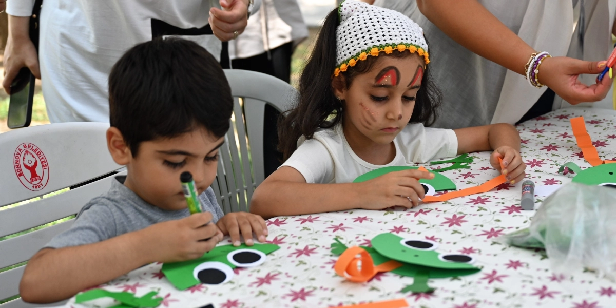 Bornova Belediyesi logolu beyaz plastik sandalyelerde oturan iki çocuk, çiçek desenli bir masa örtüsü üzerinde el işi etkinliği yapıyor. Sol taraftaki erkek çocuk elindeki kalemle yeşil renkli kağıttan kurbağa figürünü boyarken, sağ taraftaki başında beyaz örgü bir bandana bulunan ve yüzü boyalı kız çocuğu turuncu kağıt şeritlerle kurbağa figürünü süslüyor. Çocukların arkasında onlara yardımcı olan yetişkinlerin elleri ve kolları görünüyor.