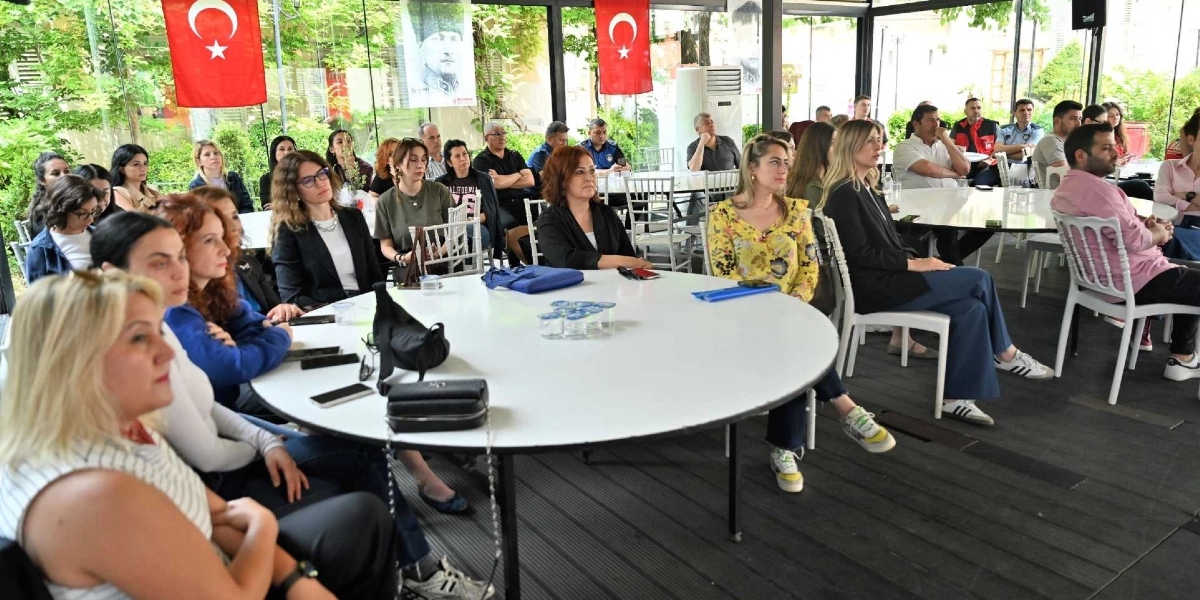 Cam duvarlarla çevrili aydınlık bir salonda, beyaz yuvarlak masaların etrafında oturmuş, dikkatle dinleyen bir grup insan. Arka planda asılı Türk bayrakları ve Mustafa Kemal Atatürk'ün bir portresi yer alıyor. Katılımcılar, afet bilinci konulu bir toplantı veya seminer için bir araya gelmiş görünüyor.